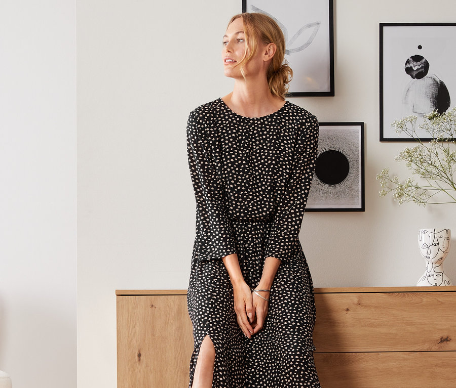 Eine Frau, die sich an eine Holzkommode lehnt, trägt eine Bluse mit Print.