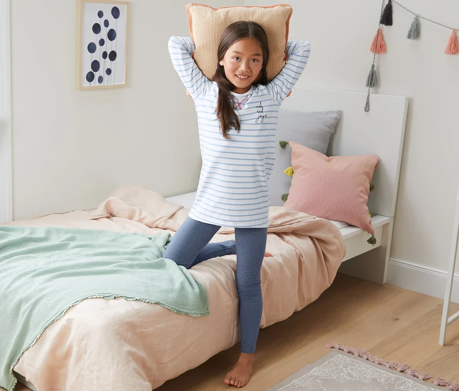 Mädchen im Mädchen-Pyjama steht am Bett und hält ein Kissen über den Kopf.