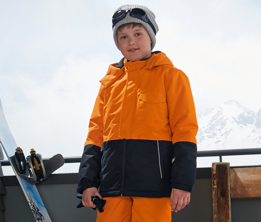 Ein Junge in einer Ski- und Snowboardjacke, Skihose, Mütze und Brille steht draußen mit Bergen im Hintergrund.