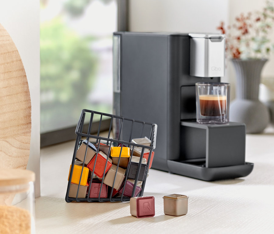 Kaffeemaschine mit Tasse und schwarzem Kapselkorb voller Kapseln steht auf einem Tisch.