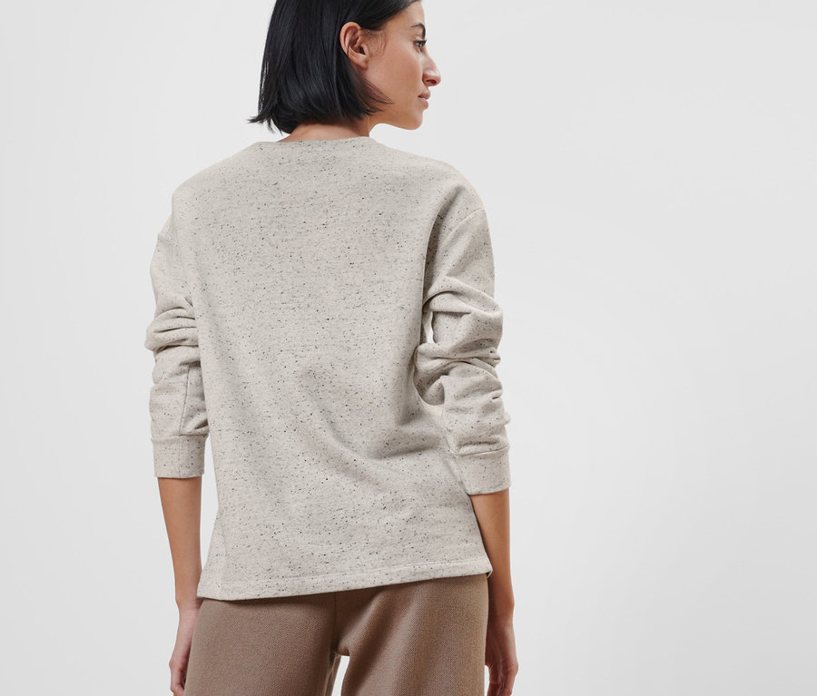 Frau steht mit dem Rücken zur Kamera und trägt ein Sweatshirt mit Knopfleiste und eine braune Hose.