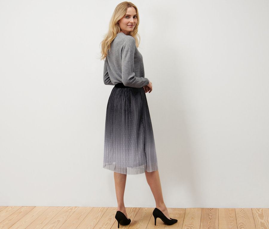 Frau posiert und trägt einen plissierten Tüllrock mit Farbverlauf, einen grauen Pullover und schwarze High Heels.