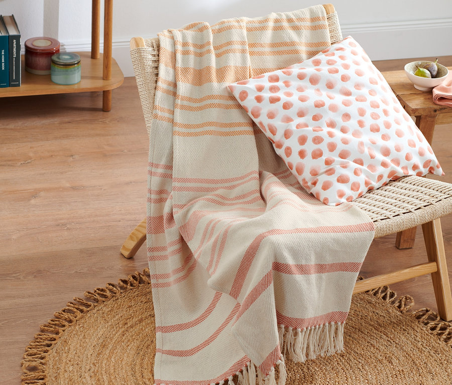 Ein Rattansessel mit einer beige Wohndecke mit orangefarbenen Streifen und einem Kissen mit orangefarbenen Punkten auf einem runden Jute-Teppich.