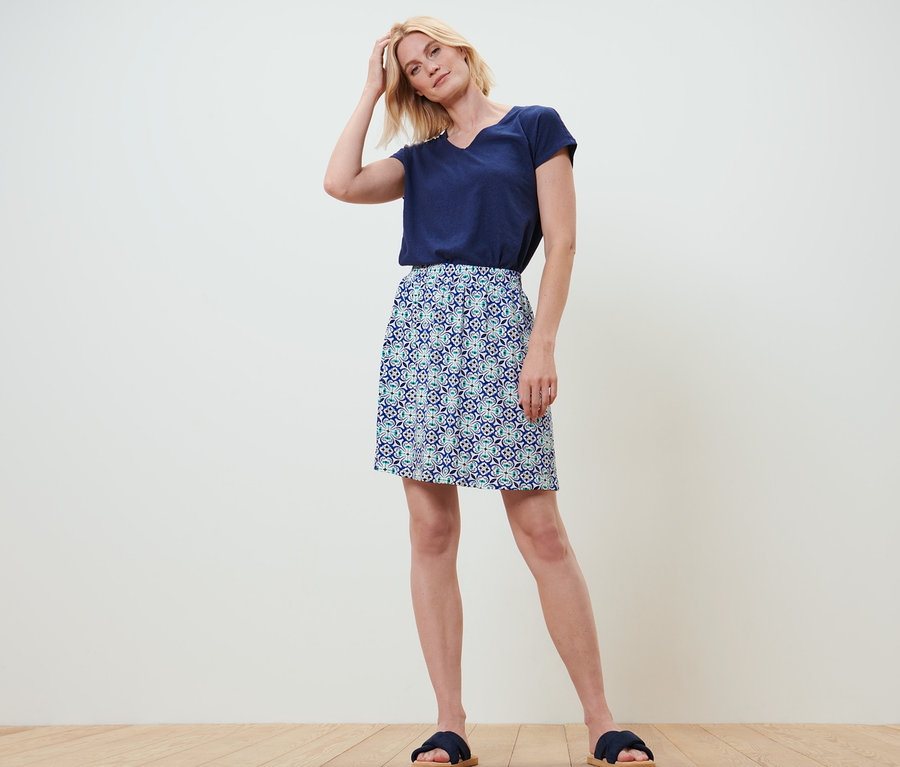 Blonde Frau steht mit der Hand im Haar. Sie trägt ein marineblaues T-Shirt und einen Jerseyrock mit Alloverprint.