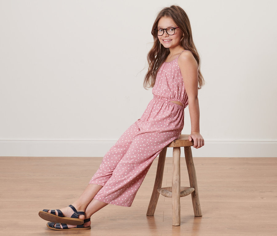 Mädchen sitzt auf einem Hocker und trägt einen Kinder-Web-Jumpsuit in 7/8-Länge.