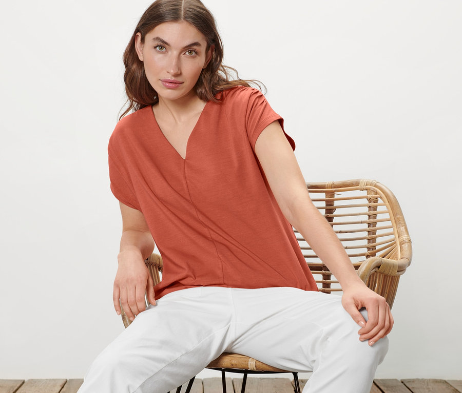 Frau sitzt auf einem Rattansessel und trägt ein apricotfarbenes Blusenshirt und eine weiße Hose.