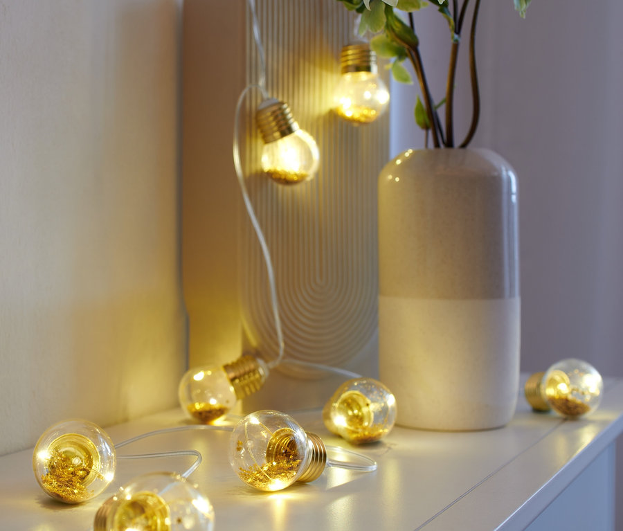 Eine LED-Glühbirnen-Lichterkette liegt auf einem weißen Regal neben einer Vase und einem Bilderrahmen.