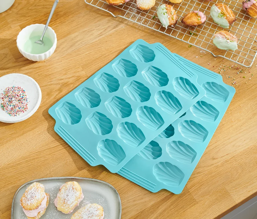 Auf einem Holztisch liegen 2 Madeleine-Backformen, gebackene Madeleines, Zuckerstreusel und Glasuren.