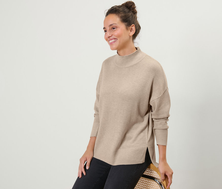 Eine lächelnde Frau sitzt und trägt einen beigen Feinstrickpullover mit Stehkragen und eine schwarze Jeans.