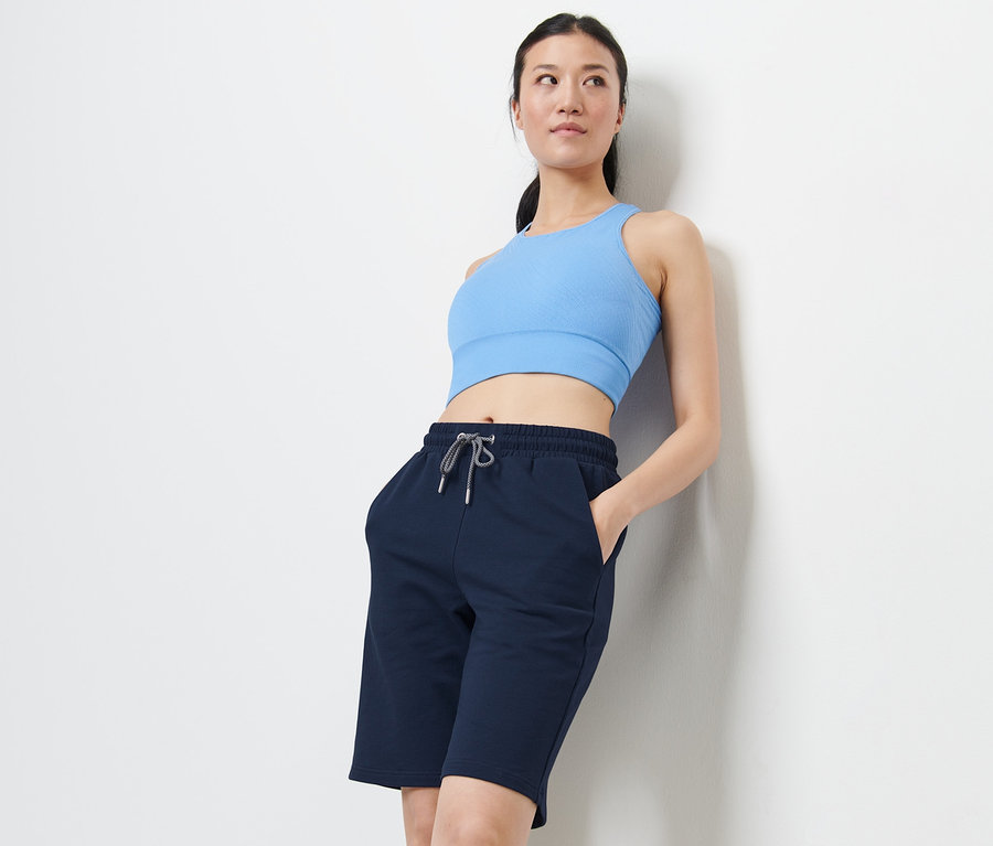 Eine Frau lehnt an einer Wand und trägt ein blaues Sportoberteil und Sportshorts.
