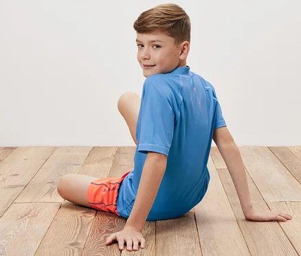 Junge sitzt auf Holzboden und trägt ein blaues Kinder-Shirt mit UV-Schutzfaktor 80 und orangefarbene Shorts.