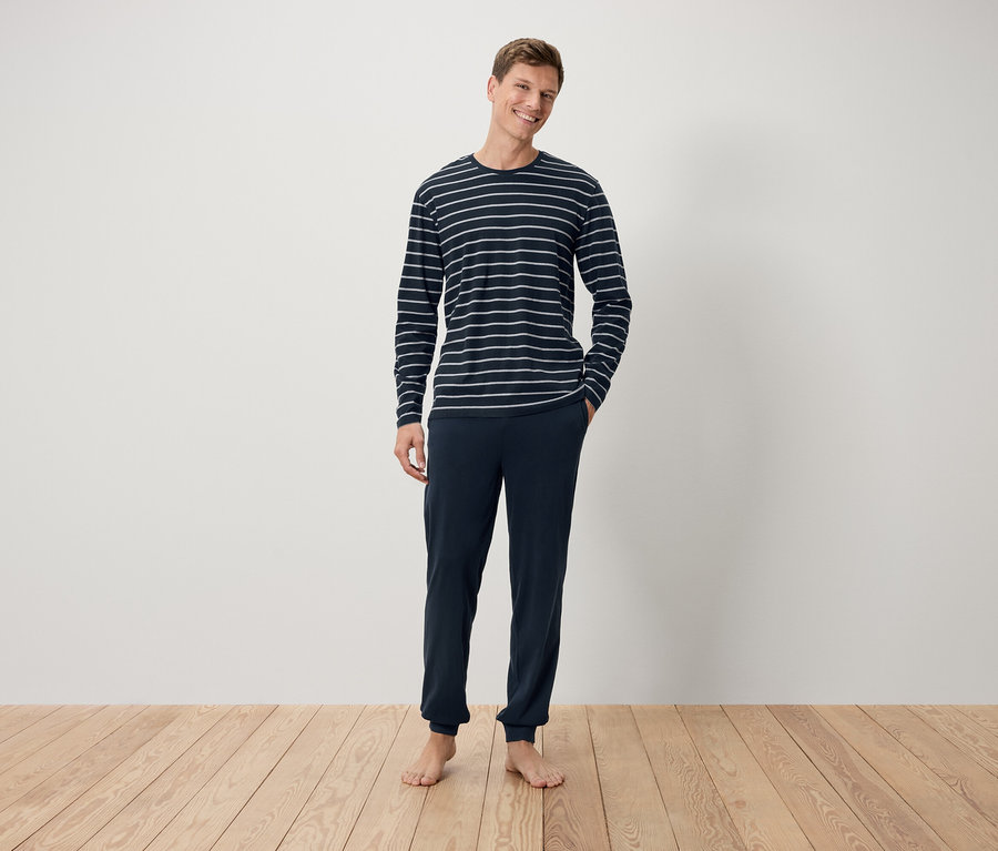 Mann steht barfuß auf einem Holzboden und trägt ein dunkelblaues, gestreiftes Langarmshirt und eine dunkelblaue Hose.