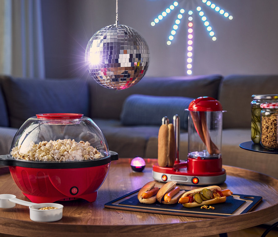 Auf einem Tisch stehen eine rote Popcornmaschine, ein Hot-Dog-Zubereiter und Hotdogs mit Gurken, im Hintergrund eine Discokugel.