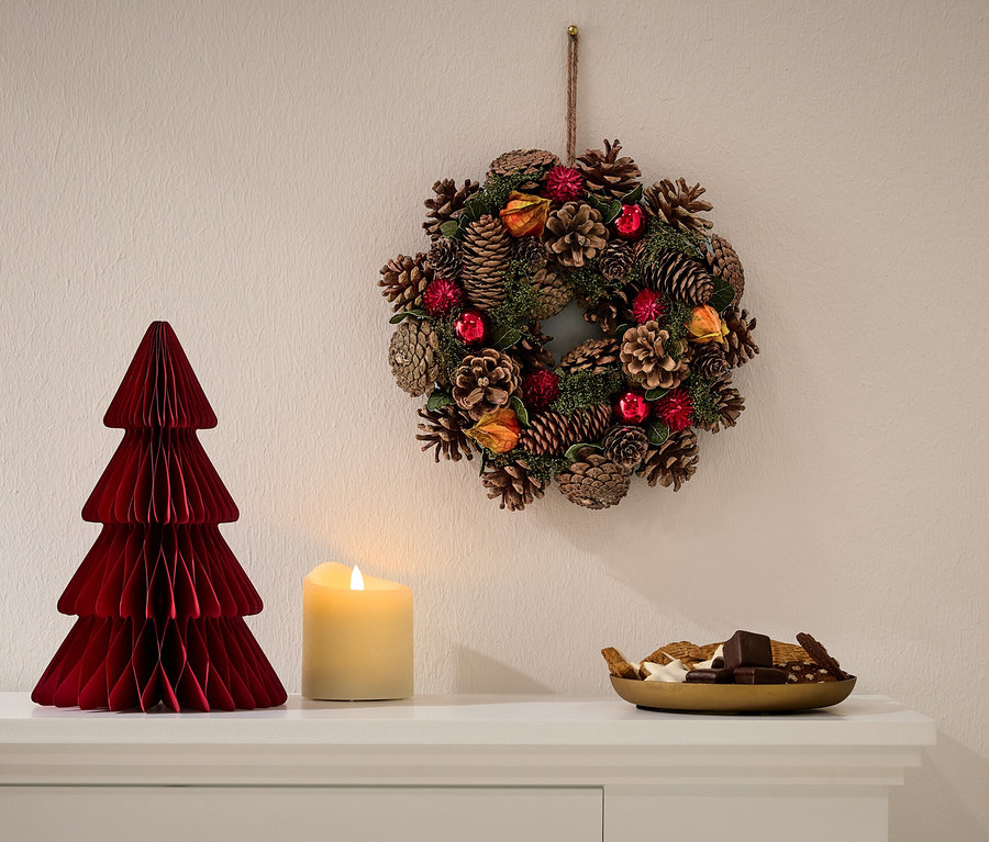 Auf einer weißen Kommode stehen ein roter Papierweihnachtsbaum, eine Kerze und eine Schale mit Süßigkeiten. An der Wand hängt ein Weihnachtskranz.