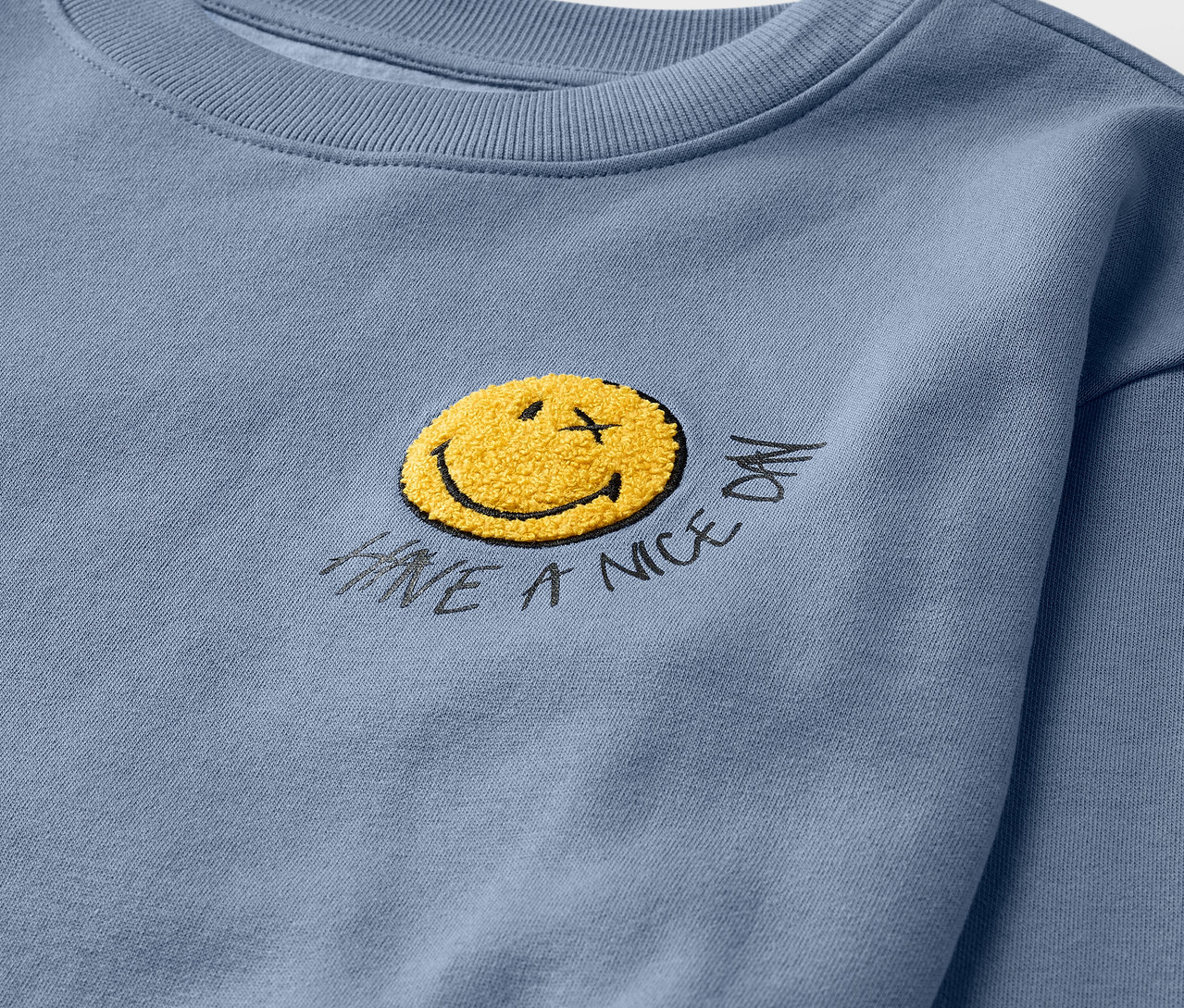 Detailaufnahme eines hellblauen Kinder-Sweatshirts SMILEYWORLD® mit angerauter Innenseite und gelbem Smiley.