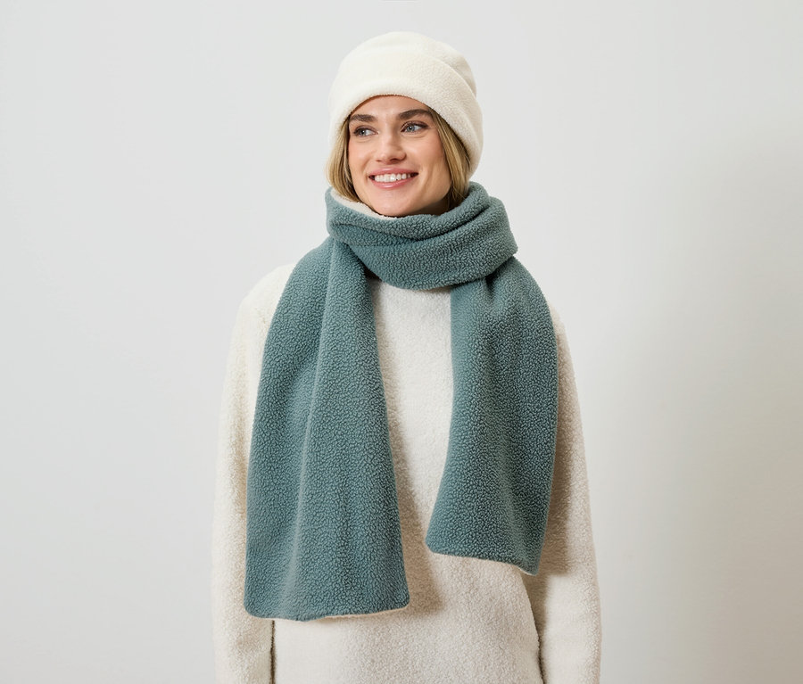 Eine lächelnde blonde Frau trägt einen cremefarbenen Pullover, einen blaugrünen Schal und eine cremefarbene Mütze.
