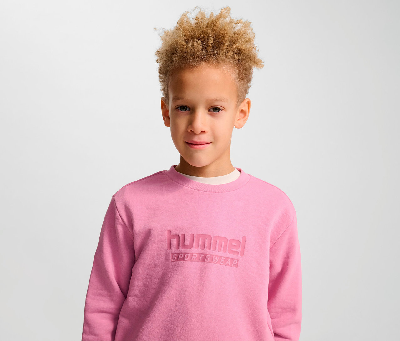 Porträt eines Jungen mit lockigen blonden Haaren in einem rosa HUMMEL HMLJR Base-Sweatshirt.