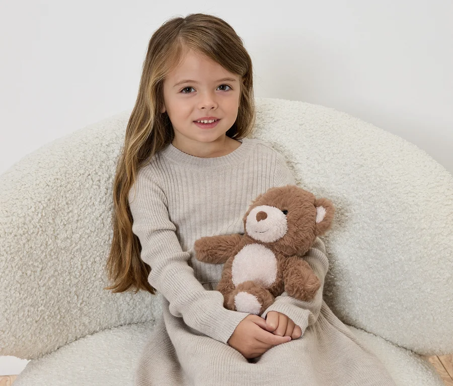 Mädchen in beigem Kleid sitzt auf einem Sessel und umarmt einen braunen Teddybären.