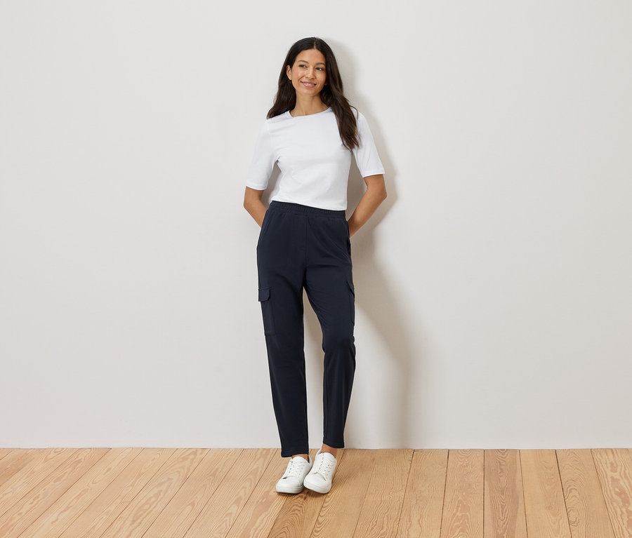 Frau steht mit verschränkten Händen vor einer Wand, trägt ein weißes T-Shirt, marineblaue Cargohose und weiße Turnschuhe.