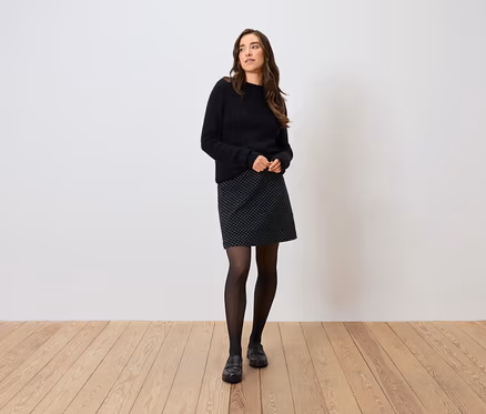 Frau posiert in schwarzem Pullover, gepunktetem Rock, schwarzen Strumpfhosen und Schuhen.