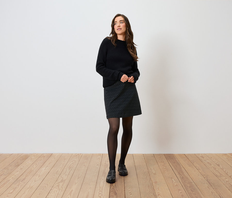 Frau posiert in schwarzem Pullover, gepunktetem Rock, schwarzen Strumpfhosen und Schuhen.
