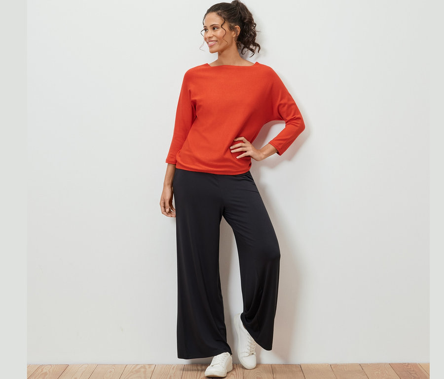 Frau posiert in orangefarbenem Feinstrickpullover, schwarzen Hosen und weißen Turnschuhen.