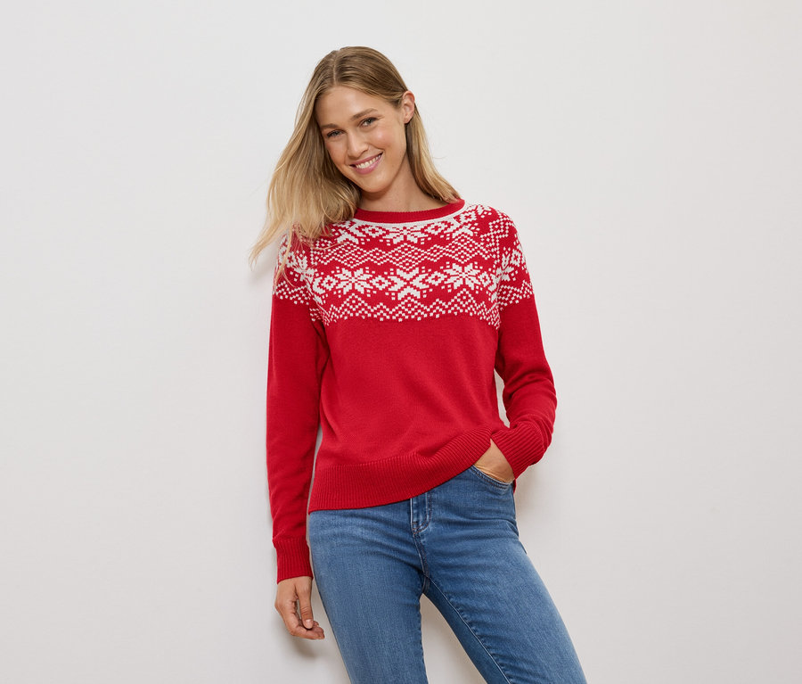 Frau mit blonden Haaren posiert in einem roten Pullover mit Schneeflockenmuster und blauen Jeans.