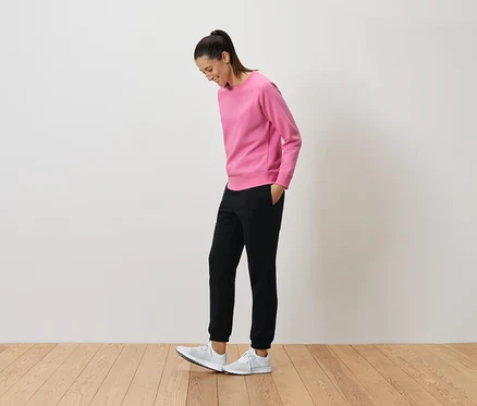 Frau in pinkem Pullover und schwarzen Hosen schaut auf ihre weißen Turnschuhe.