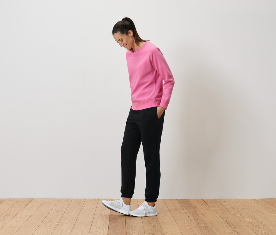 Frau in pinkem Pullover und schwarzen Hosen schaut auf ihre weißen Turnschuhe.