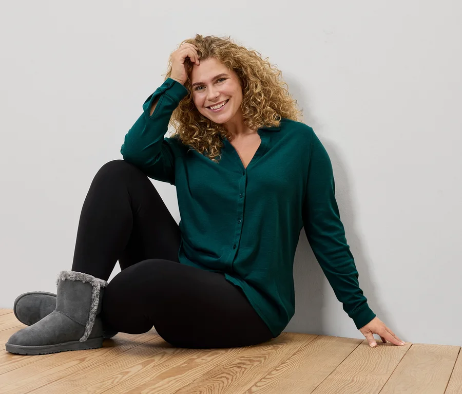 Frau sitzt auf dem Boden, trägt ein grünes Hemd, schwarze Leggings und graue Stiefel.