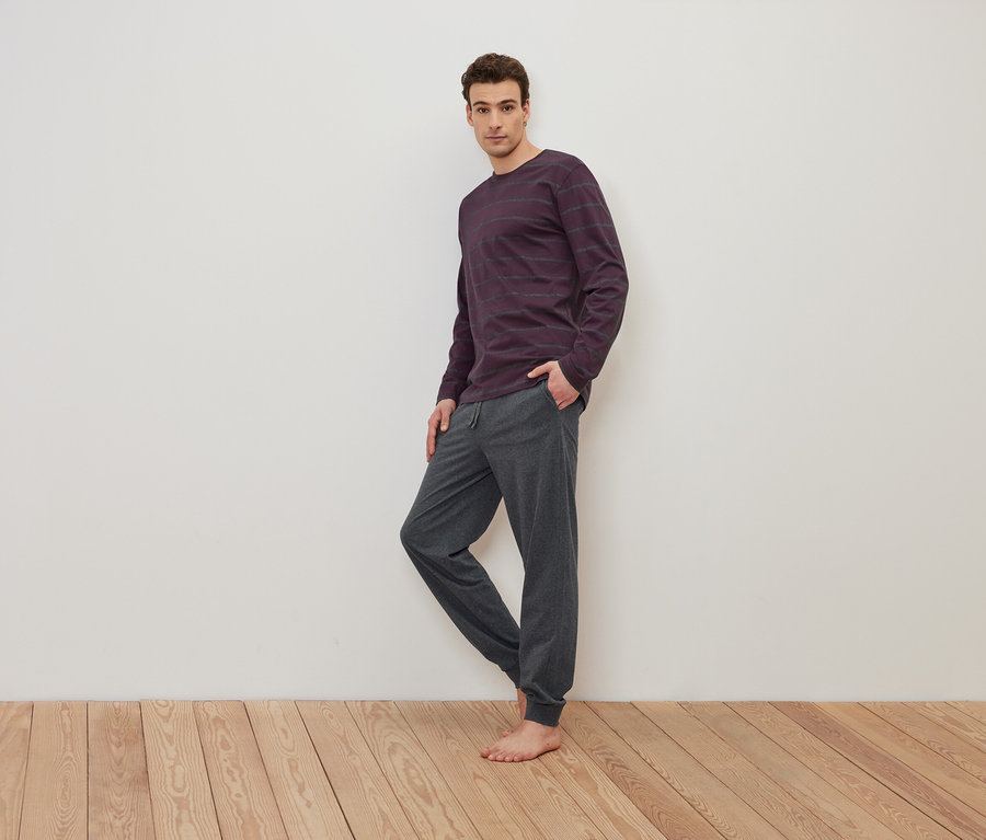 Ein Mann steht barfuß auf einem Holzboden und trägt ein lila gestreiftes Langarmshirt und eine graue Jogginghose.