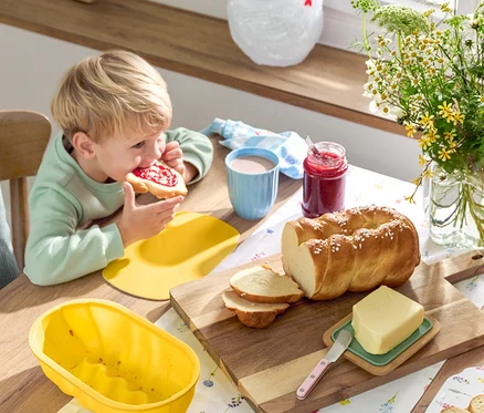 Blonder Junge isst ein Marmeladenbrot am Tisch mit Striezel, Butter, Marmelade, Geschirr und Blumen.