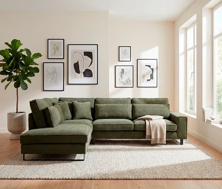 Grünes Ecksofa mit Kissen und Decke auf einem Teppich. An der Wand hängen sechs gerahmte Bilder.