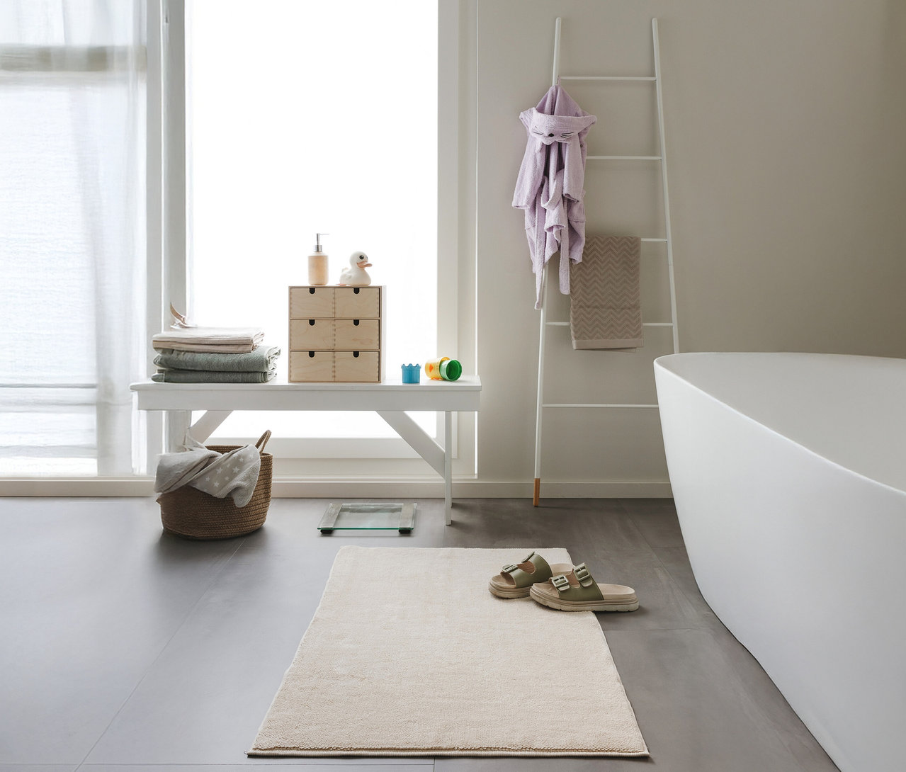 Ein beiges Badezimmer mit einem weißen Hocker, auf dem gefaltete Handtücher und eine Kommode stehen, sowie einer beigen Badematte »Ole« von benuta (50 x 80 cm).