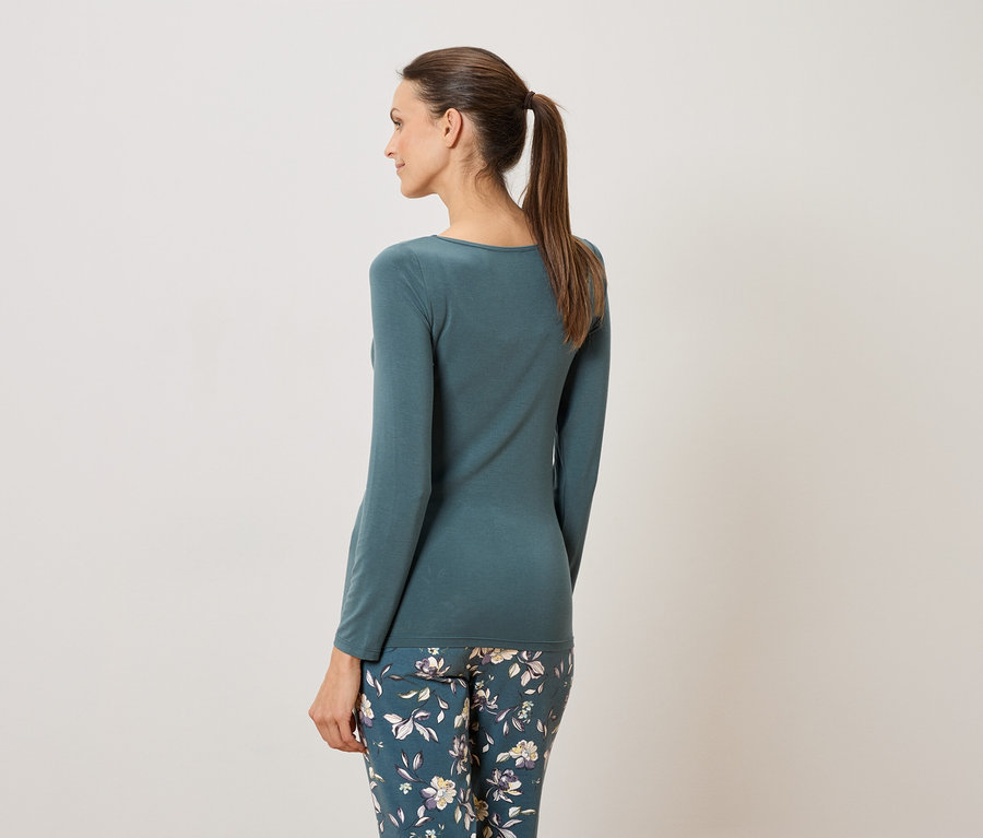 Frau von hinten mit blaugrünem, langärmligem Oberteil und blumiger Hose.