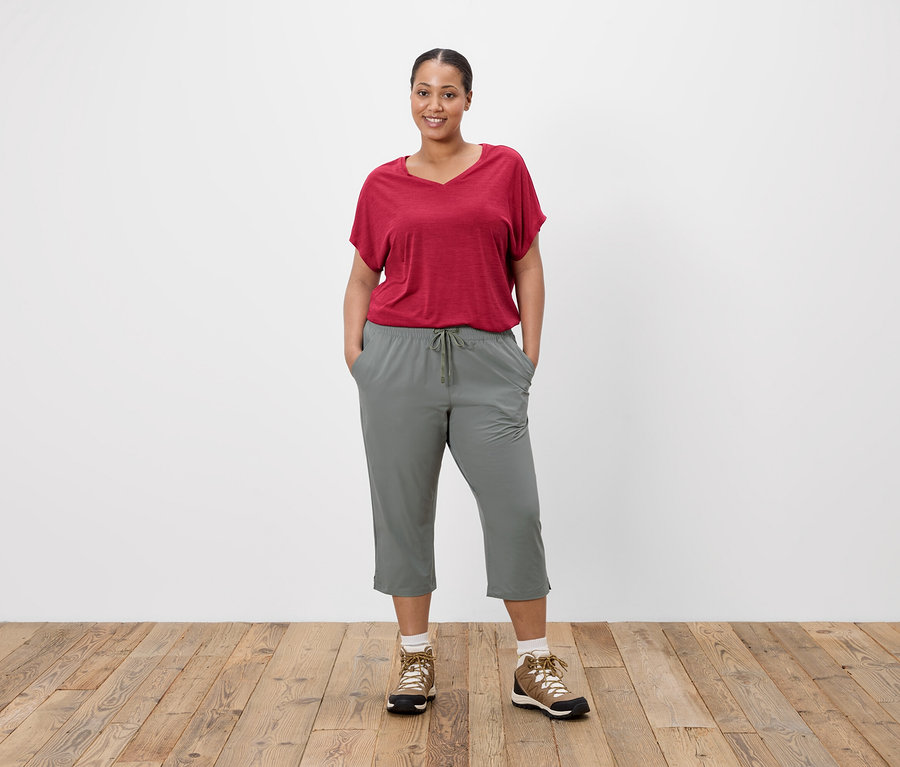 Frau mit rotem T-Shirt und grauer Hose, die vor einer weißen Wand steht.