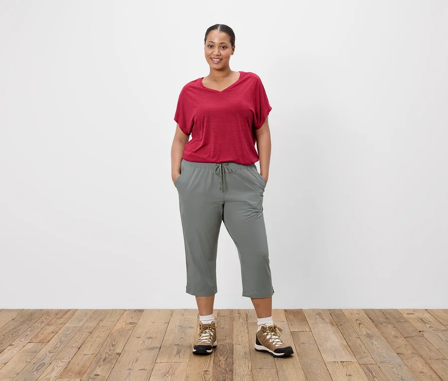 Frau mit rotem T-Shirt und grauer Hose, die vor einer weißen Wand steht.