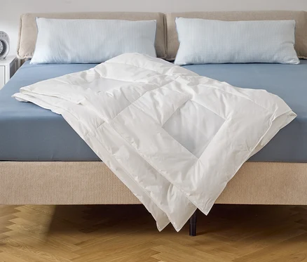 Eine weiße Bettdecke liegt auf einem Bett mit blauem Spannbettlaken und zwei Kissen. Auf dem Nachttisch steht ein Ventilator.