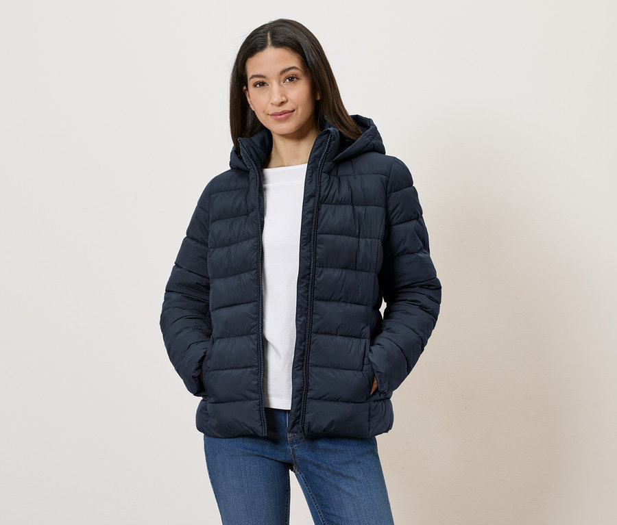 Frau mit marineblauer Steppjacke mit Kapuze, weißem T-Shirt und Jeans, die mit den Händen in den Taschen vor einer weißen Wand steht.