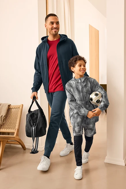 Mann mit Sporttasche und Kind mit Fußball, beide tragen Sportkleidung. Der Mann trägt eine Sweatjacke und Sweathose. Das Kind trägt eine Kinder-Windbreaker-Jacke.