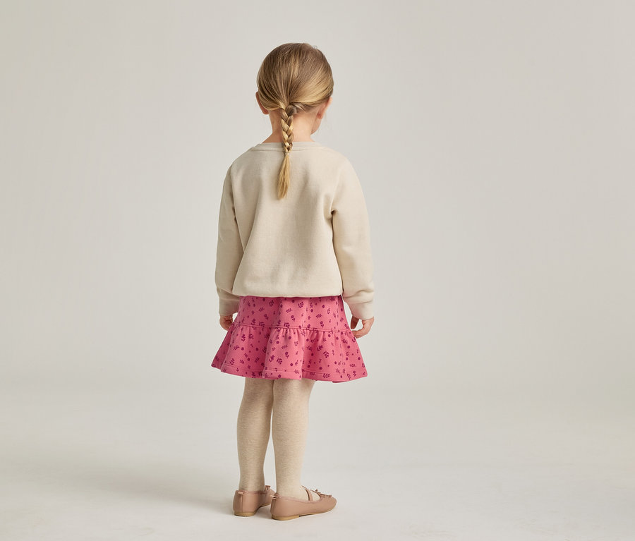 Rückansicht eines Mädchens mit blondem Zopf, das einen beigen Pullover, einen rosa Rock mit Rüschen und Ballerinaschuhe trägt.