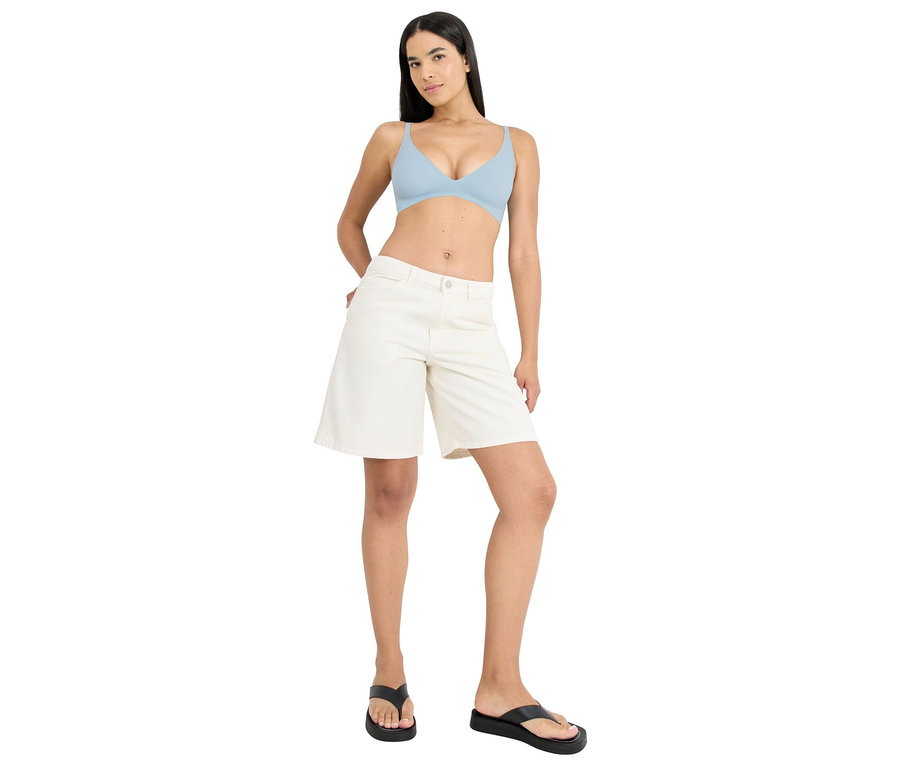 Frau posiert in hellblauem BH, cremefarbenen Shorts und schwarzen Flip-Flops, Hände in den Hosentaschen.