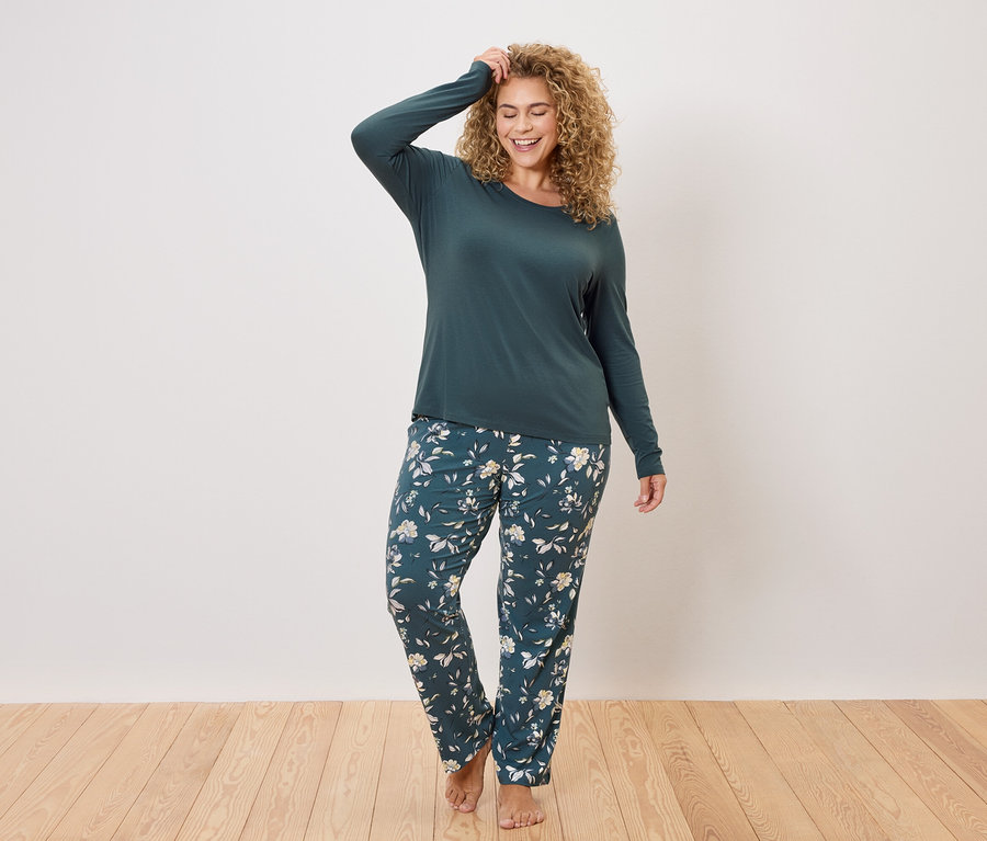 Frau mit lockigem Haar trägt ein dunkelgrünes, langärmeliges Oberteil und eine dunkelgrüne, blumengemusterte Pyjamahose.