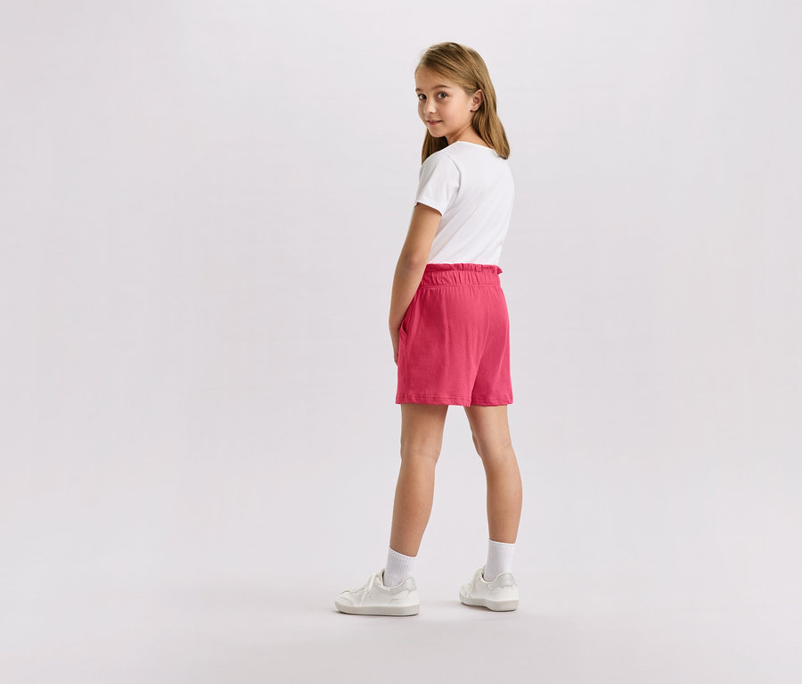 Mädchen mit blonden Haaren, das ein weißes T-Shirt, rosafarbene Shorts und weiße Turnschuhe trägt und in die Kamera blickt.