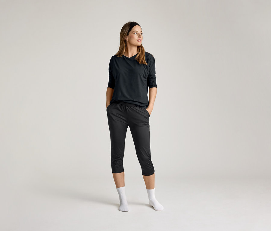 Frau mit schwarzem 3/4-Sportshirt und dunkler Hose, Hände in den Taschen, weiße Socken, vor hellem Hintergrund.