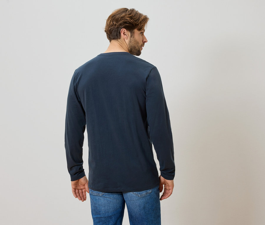 Mann von hinten in blauem Langarmshirt und Jeans vor hellem Hintergrund.