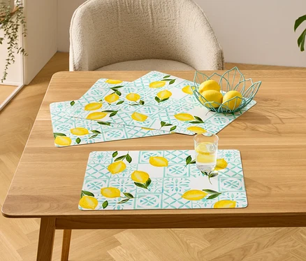 Auf einem Holztisch liegen drei Tischsets mit Zitronenmuster, eine Schale mit Zitronen und ein Glas Wasser mit Zitrone.