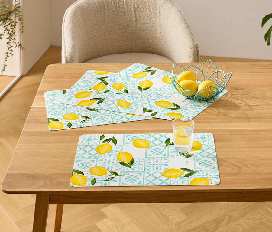 Auf einem Holztisch liegen drei Tischsets mit Zitronenmuster, eine Schale mit Zitronen und ein Glas Wasser mit Zitrone.