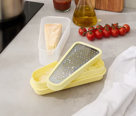 Auf einer Küchentheke stehen ein Topf, Tomaten am Zweig, eine Ölflasche, eine Schüssel mit Sauce, ein Behälter mit Käse und eine gelbe Reibe.