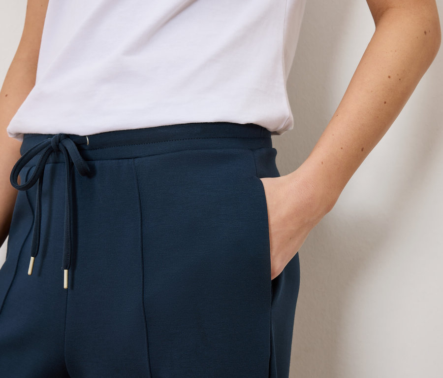 Nahaufnahme einer Frau, die marineblaue Trainingshose mit Kordelzug und goldenen Details und ein weißes T-Shirt trägt.